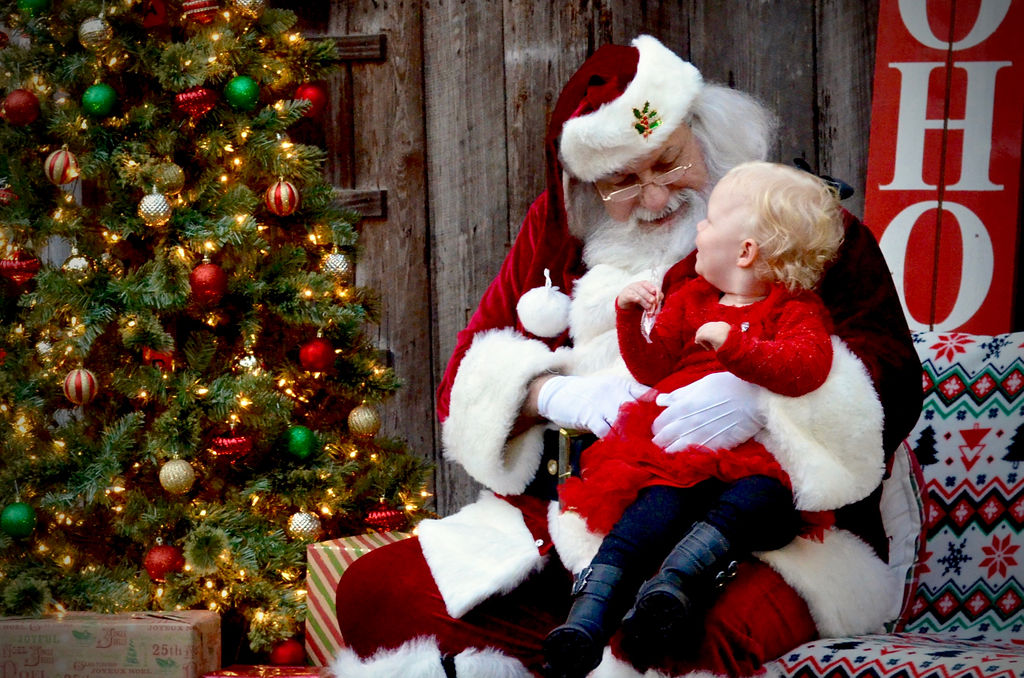 Santa Clause with a baby sitting on his lap in front of a Christmas tree.