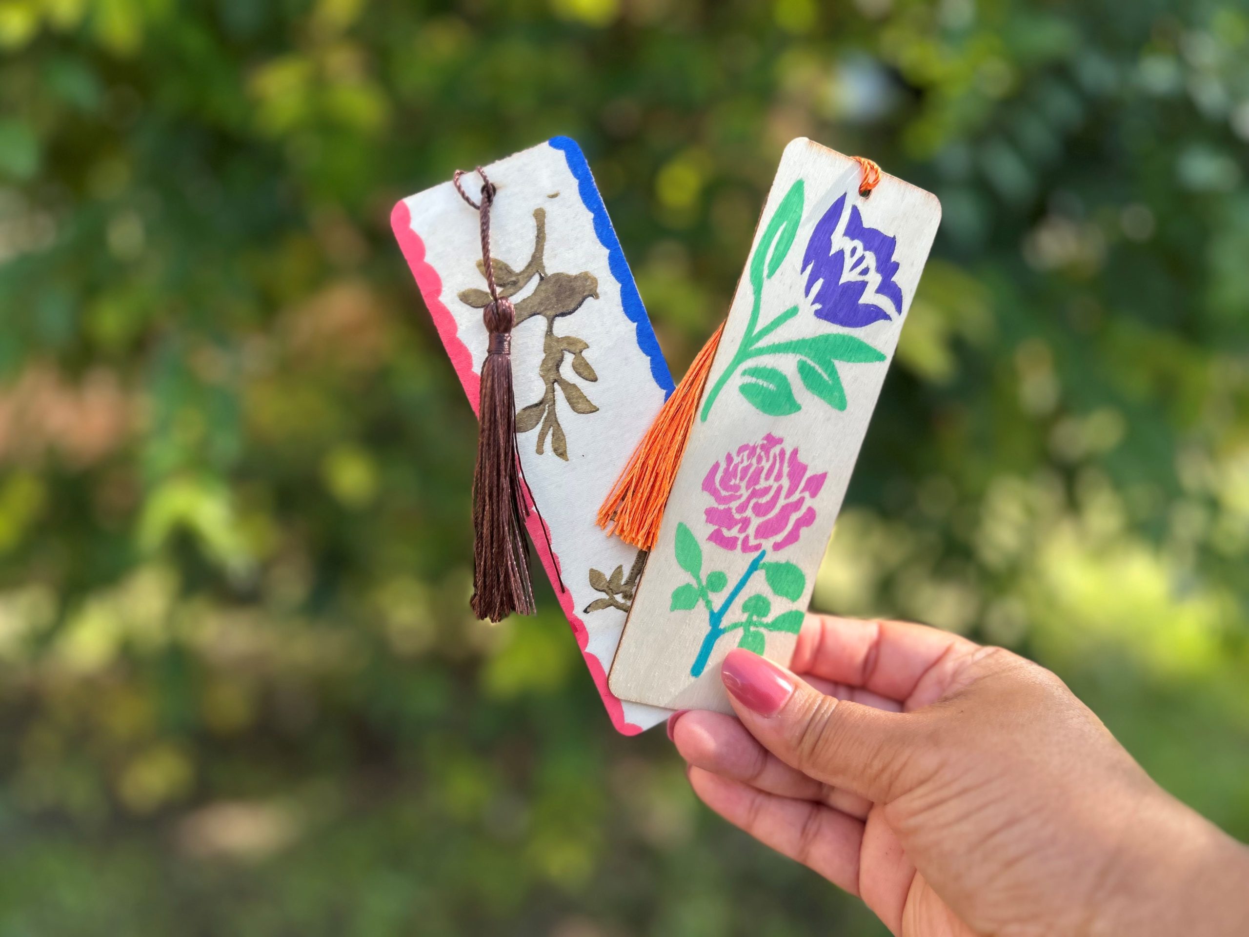 Hand holding two wooden bookmarks painted with multicolored flowers.
